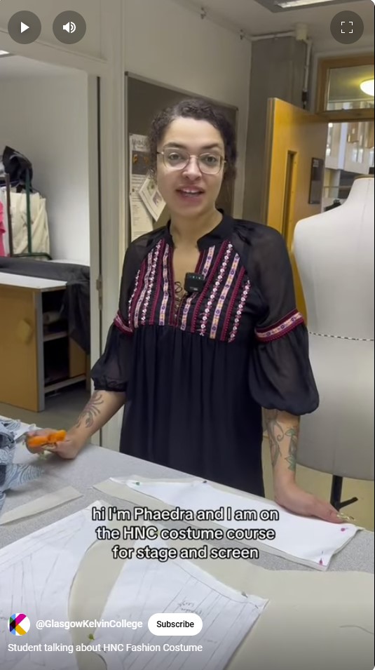 HNC costume design student presenting their work in a classroom, with sewing patterns and fabric samples on the table.