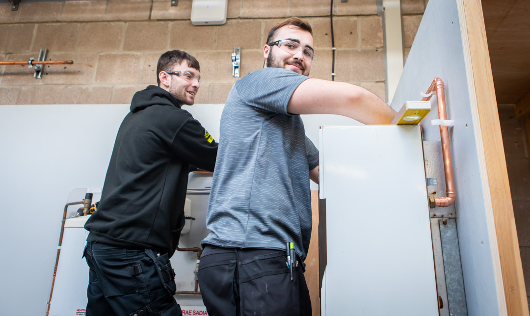 Two plumbing students smiling while collaboratively working on installing a plumbing fixture, engaged in hands-on training.