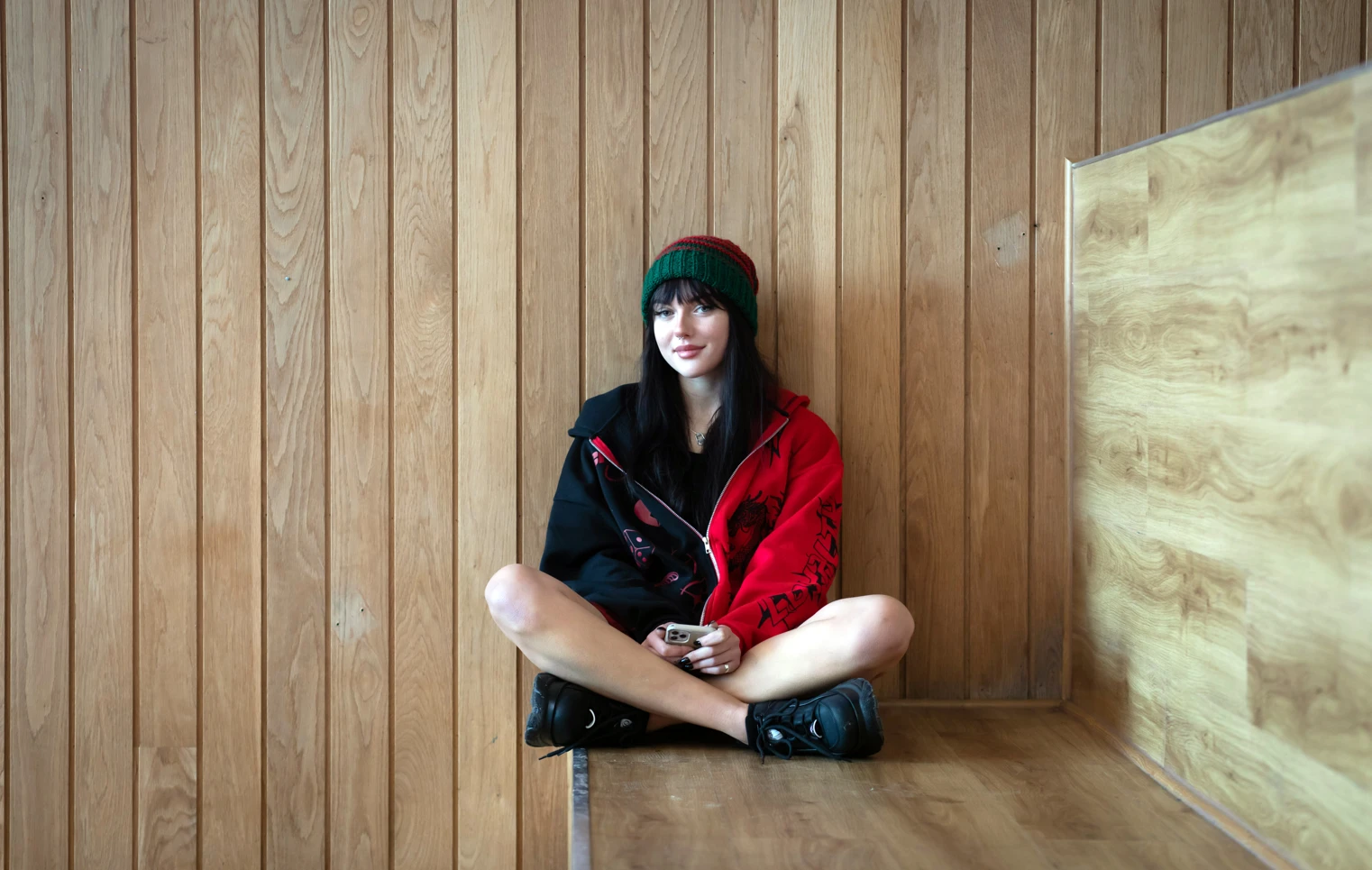 Student sitting cross-legged on a wooden bench against a wood-panelled wall at Glasgow Kelvin College’s Springburn Campus, wearing a red and black jacket and green hat. Student sitting cross-legged on a wooden bench against a wood-panelled wall at Glasgow Kelvin College’s Springburn Campus, wearing a red and black jacket and green hat.