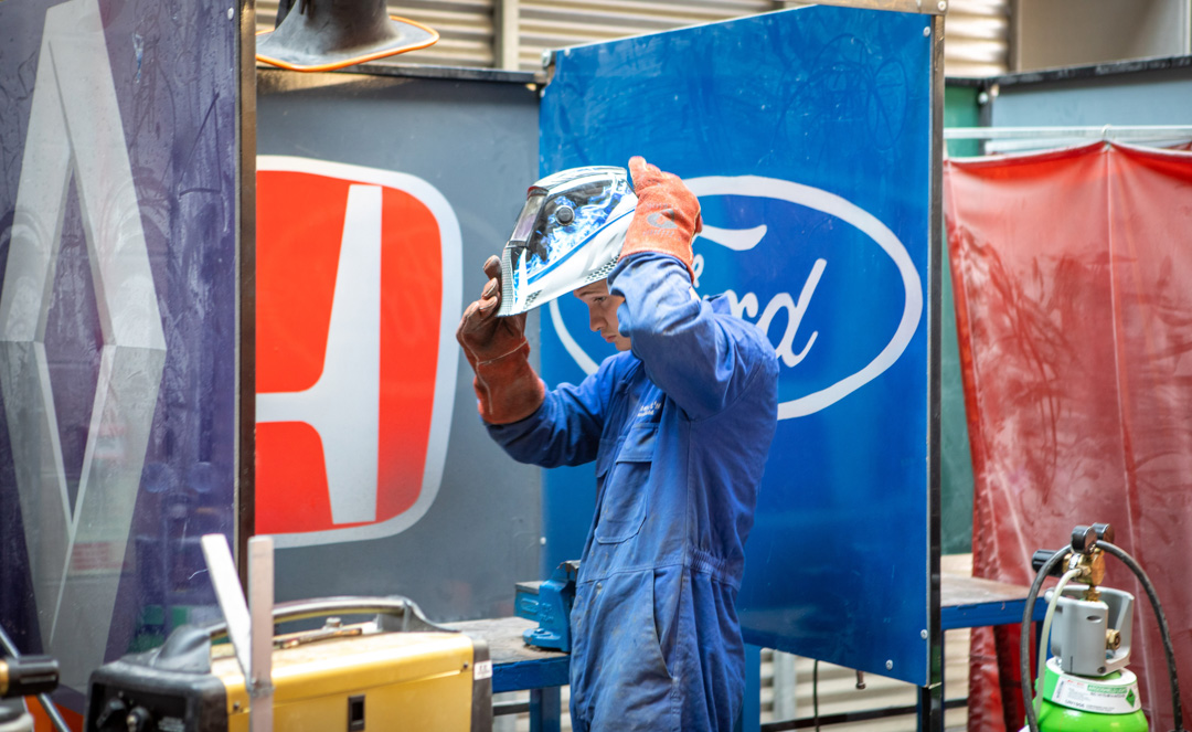 Student adjusting welding helmet beside car manufacturer screens in the automotive workshop.