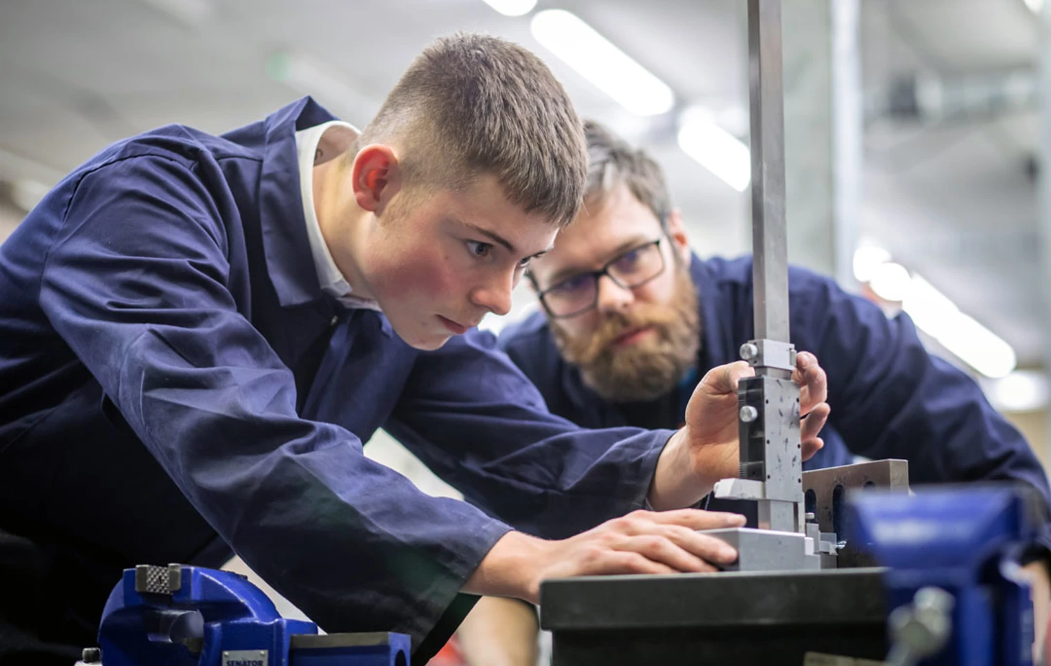A lecturer and a student in navy-blue overalls working together in a mechanical engineering workshop. The lecturer is demonstrating how to use a tool on a metal component held in a vise. A lecturer and a student in navy-blue overalls working together in a mechanical engineering workshop. The lecturer is demonstrating how to use a tool on a metal component held in a vise.