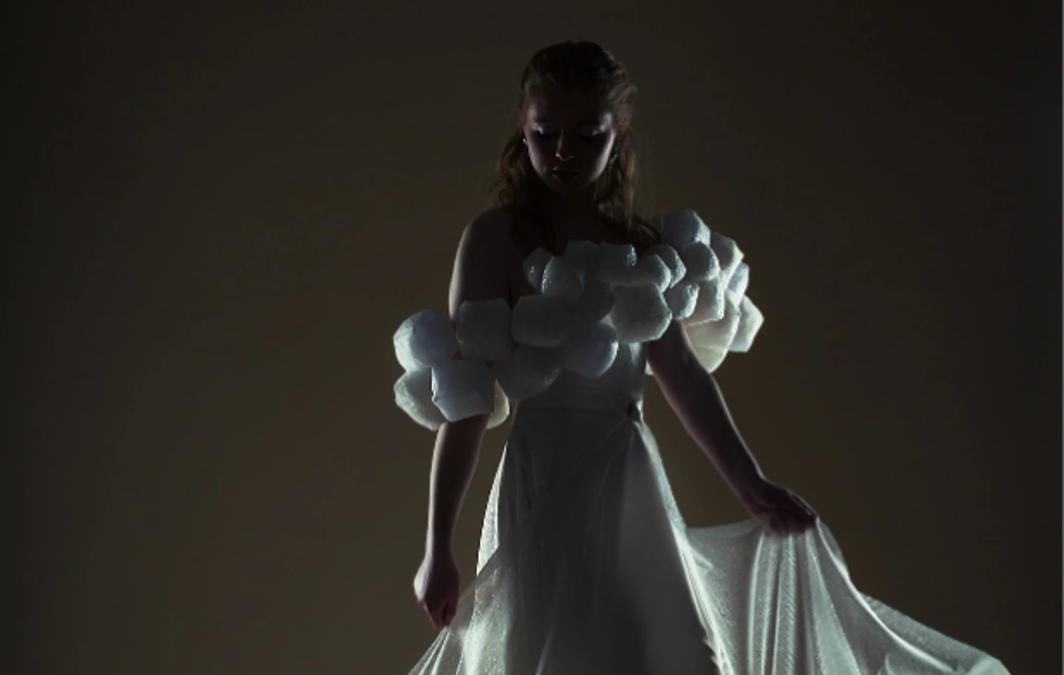 Model in a dramatic, low-lit setting wearing an off-the-shoulder white gown with textured puff sleeves and a flowing sheer train, softly illuminated from behind. Model in a dramatic, low-lit setting wearing an off-the-shoulder white gown with textured puff sleeves and a flowing sheer train, softly illuminated from behind.