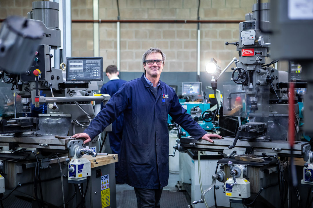 A mechanical engineering lecturer in a navy-blue coat standing confidently in a workshop, surrounded by milling machines and industrial equipment.