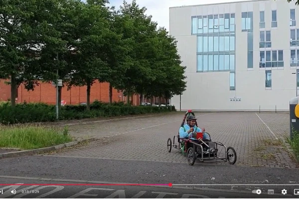 A student in a helmet and blue jacket driving a custom-built electric car in an open parking area outside a modern college building, surrounded by trees A student in a helmet and blue jacket driving a custom-built electric car in an open parking area outside a modern college building, surrounded by trees