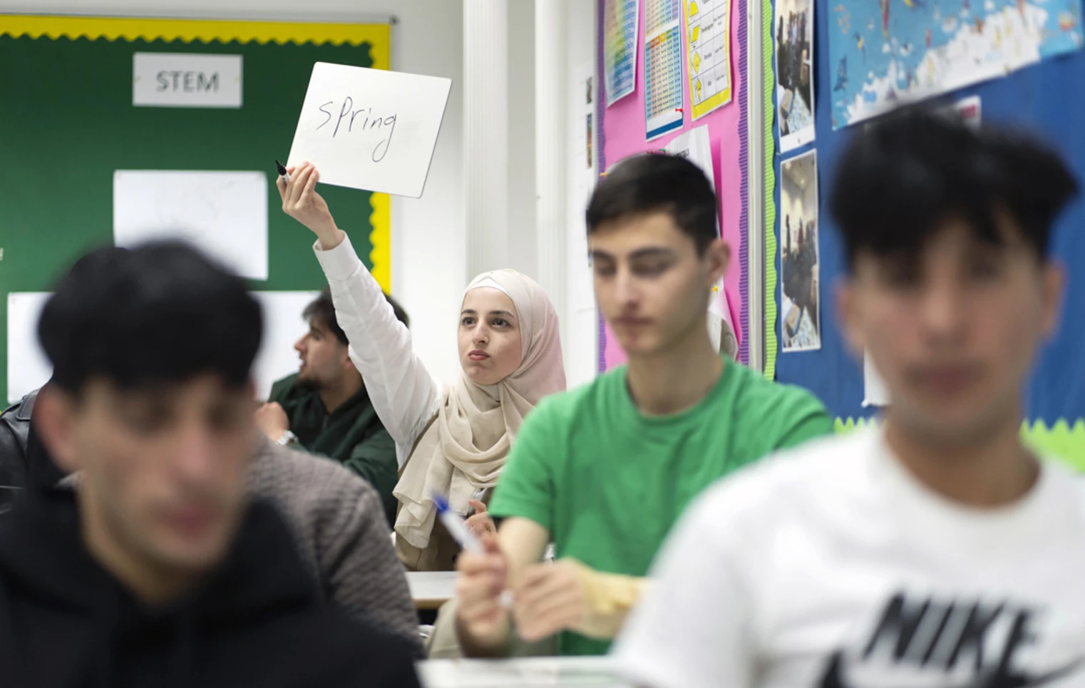 A female student in a beige hijab holds up a small whiteboard with the word "Spring" written on it, participating in a classroom activity. Other students are in the foreground, slightly out of focus. A female student in a beige hijab holds up a small whiteboard with the word "Spring" written on it, participating in a classroom activity. Other students are in the foreground, slightly out of focus.