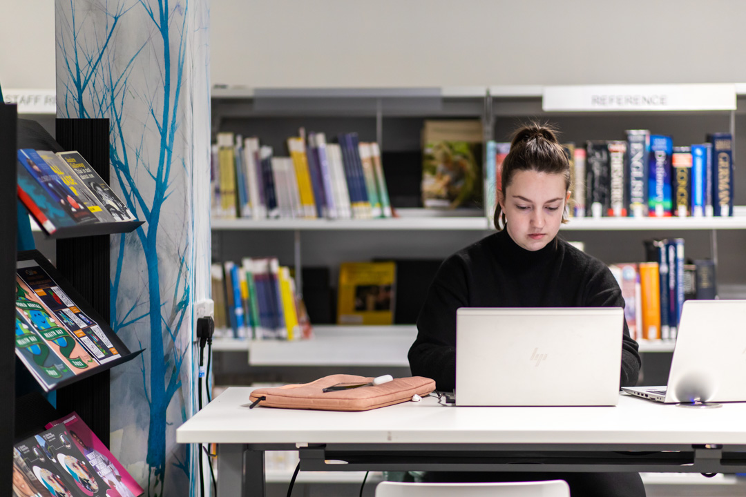 Student working on a laptop in the library at Glasgow Kelvin College’s Easterhouse campus, with bookshelves and study resources in the background.