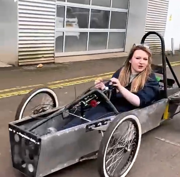 Automotive student driving a student-built race car during a practical session outside the workshop.