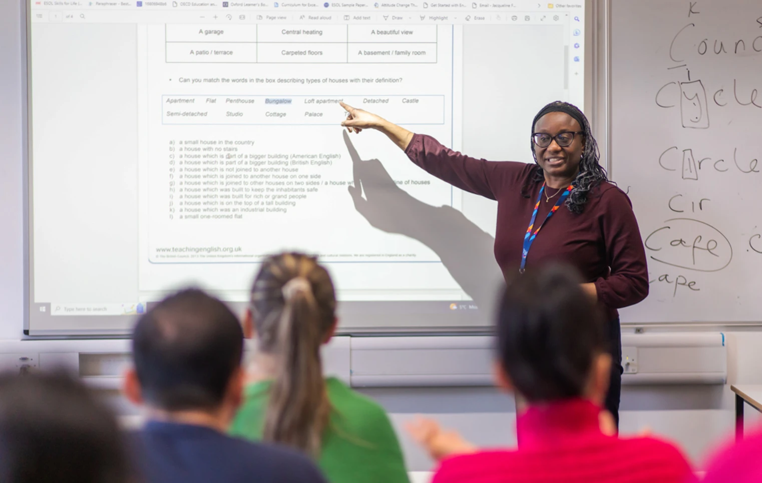 A female lecturer wearing glasses and a lanyard points to a projected lesson on a whiteboard while teaching a group of students. A female lecturer wearing glasses and a lanyard points to a projected lesson on a whiteboard while teaching a group of students.