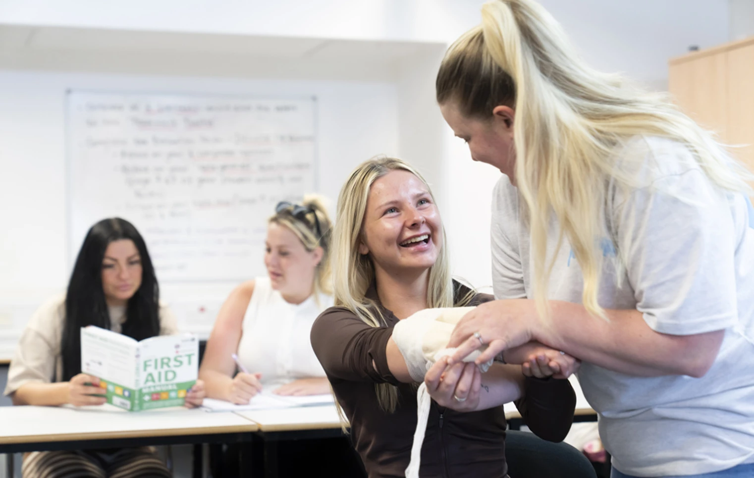A student sits at a classroom desk, holding open a first aid manual with a bandage and tablet nearby, ready to tackle some hands-on learning A student sits at a classroom desk, holding open a first aid manual with a bandage and tablet nearby, ready to tackle some hands-on learning