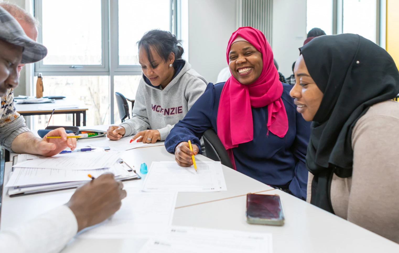 A diverse group of students, including a woman in a pink hijab, laughs and collaborates while working on an assignment in a classroom. A diverse group of students, including a woman in a pink hijab, laughs and collaborates while working on an assignment in a classroom.