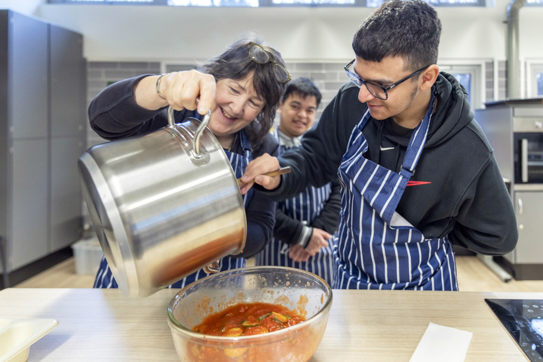 Woman pouring sauce from a large pot into a bowl while smiling and working with two young men in striped aprons. Woman pouring sauce from a large pot into a bowl while smiling and working with two young men in striped aprons.