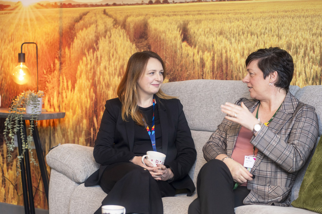 Two Glasgow Kelvin College staff members sitting on a sofa with coffee mugs, having a friendly conversation in a relaxed setting with a scenic wall backdrop.