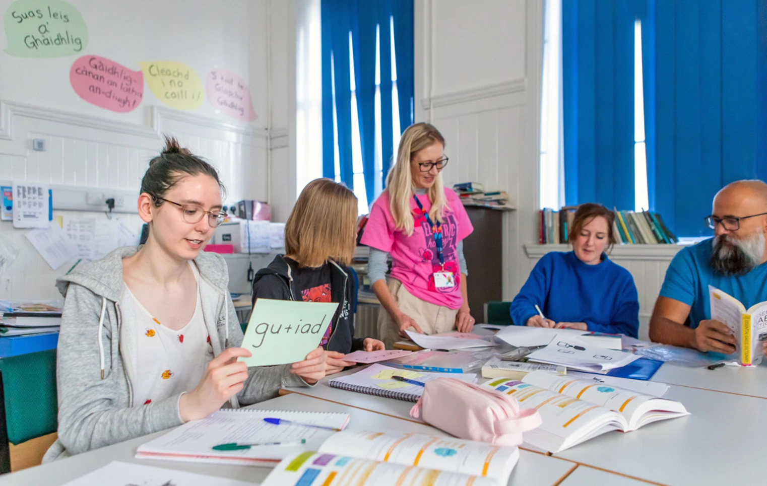 Gaelic lecturer supporting students in a language learning activity, with students holding vocabulary cards and engaging in conversation. Gaelic lecturer supporting students in a language learning activity, with students holding vocabulary cards and engaging in conversation.
