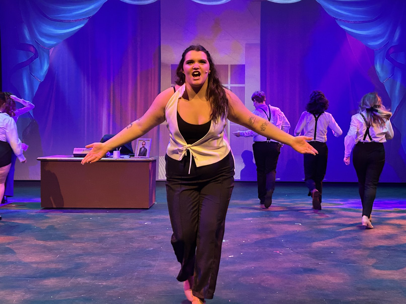 Performer delivering a powerful monologue, surrounded by other actors on stage in dramatic poses.
