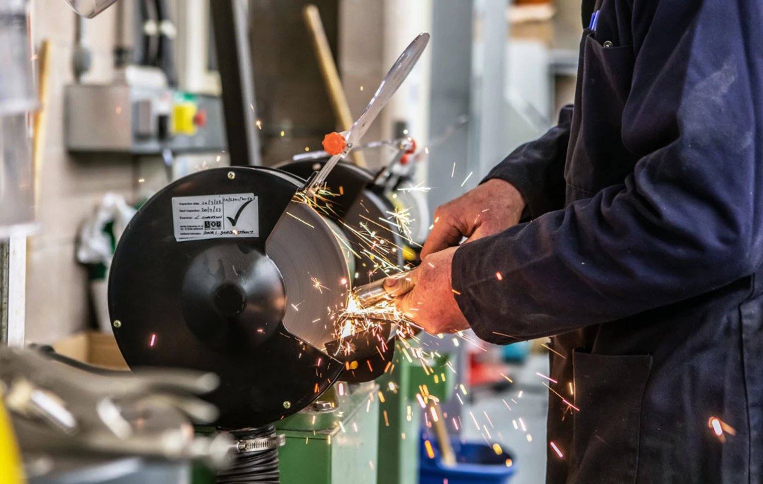 Close-up of a grinding wheel in action, with sparks flying as a lecturer in a navy-blue coat sharpens a metal component. Close-up of a grinding wheel in action, with sparks flying as a lecturer in a navy-blue coat sharpens a metal component.