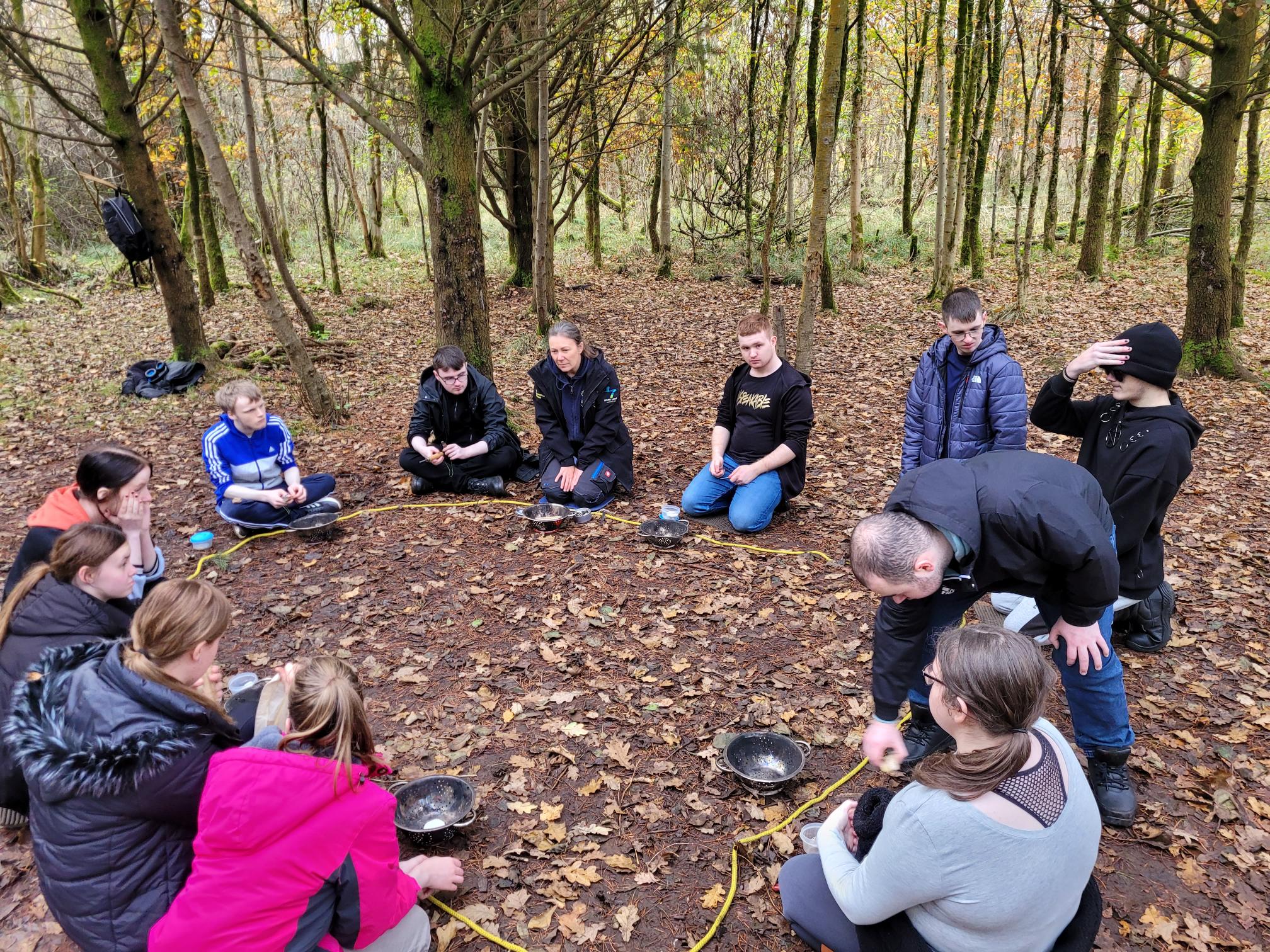 Group of young people sitting in a circle on the forest floor, engaged in a team-building or outdoor activity.