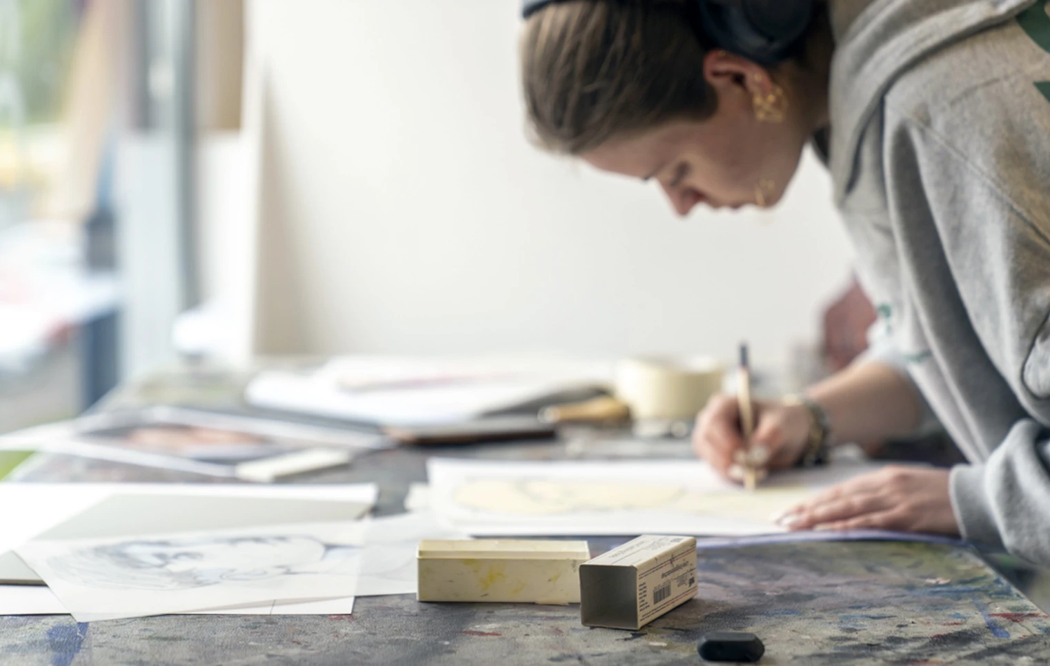 Person leaning over a table, drawing on paper in an art studio, with various sketches and art materials spread out Person leaning over a table, drawing on paper in an art studio, with various sketches and art materials spread out