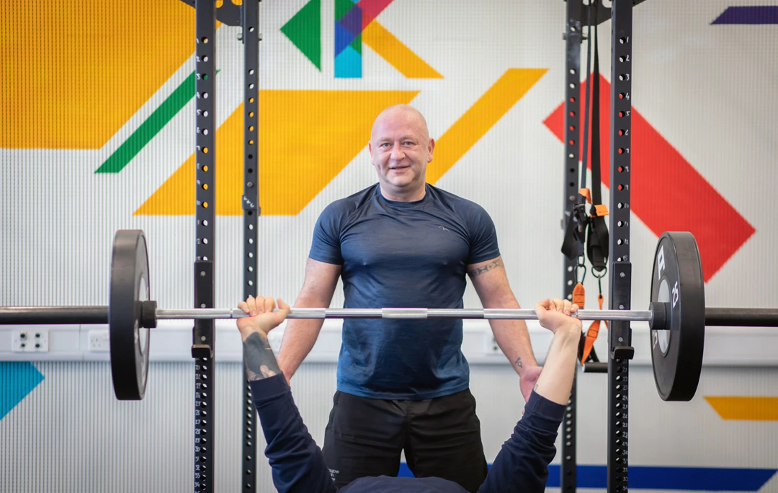 Student performing a bench press while another student spots them in a colourful training area. Student performing a bench press while another student spots them in a colourful training area.