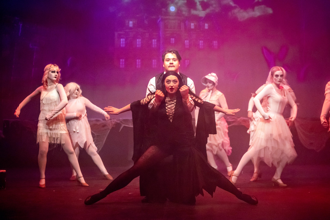 Group of musical theatre students performing a choreographed ensemble scene on stage, with central performers in dark costumes and dancers in white behind them.