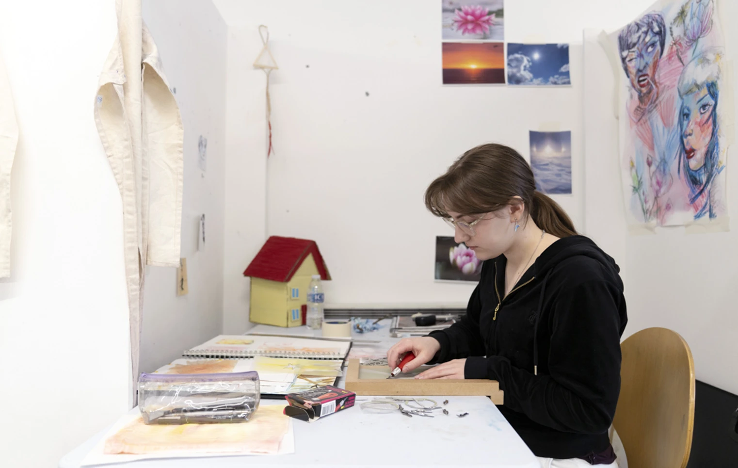 Person seated at a desk in an art studio, carving a design into a printmaking block, with sketchbooks and artwork on the wall Person seated at a desk in an art studio, carving a design into a printmaking block, with sketchbooks and artwork on the wall