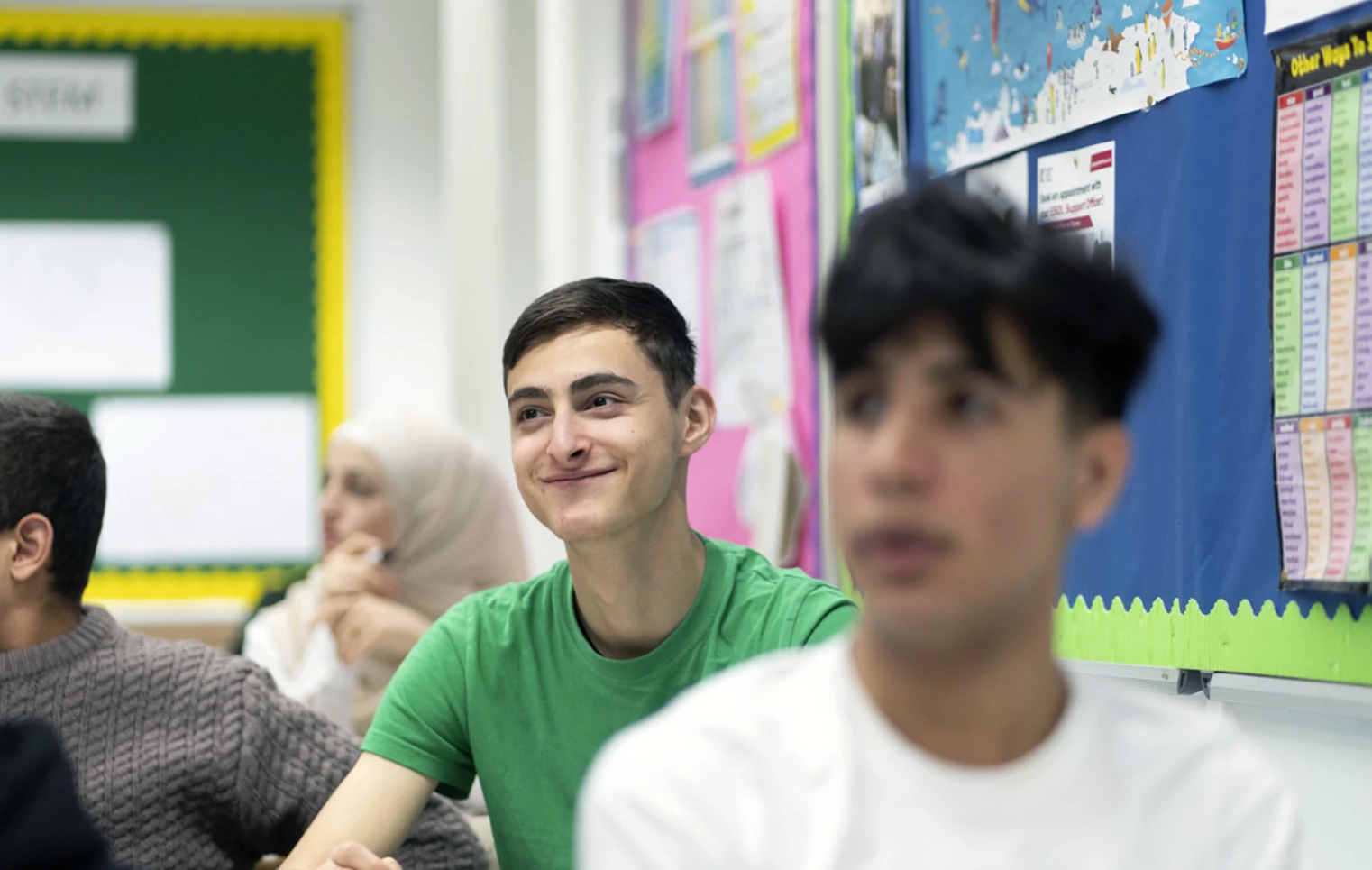 A young male student in a green t-shirt sits in a classroom, smiling while holding a whiteboard marker. Other students are visible in the background, including a student in a white hijab. A young male student in a green t-shirt sits in a classroom, smiling while holding a whiteboard marker. Other students are visible in the background, including a student in a white hijab.