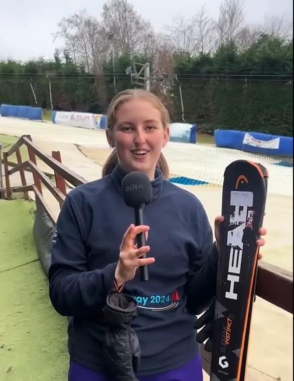 Outdoor Adventure Sports student Sarah speaking into a microphone while holding skis at an outdoor ski slope.