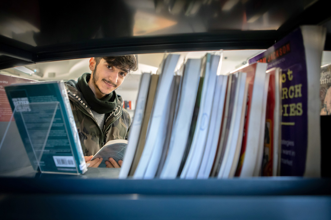 Student browsing books in the East End campus library at Glasgow Kelvin College, viewed through a shelf.