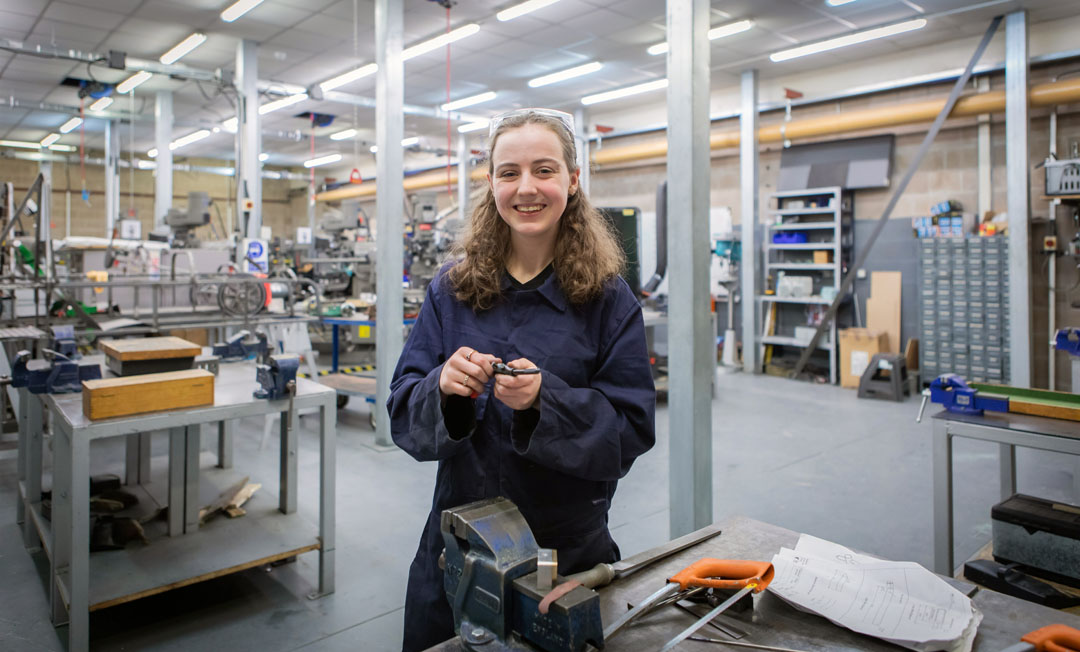 A female mechanical engineering student in navy-blue overalls smiling while working on a small metal component in a busy college workshop.