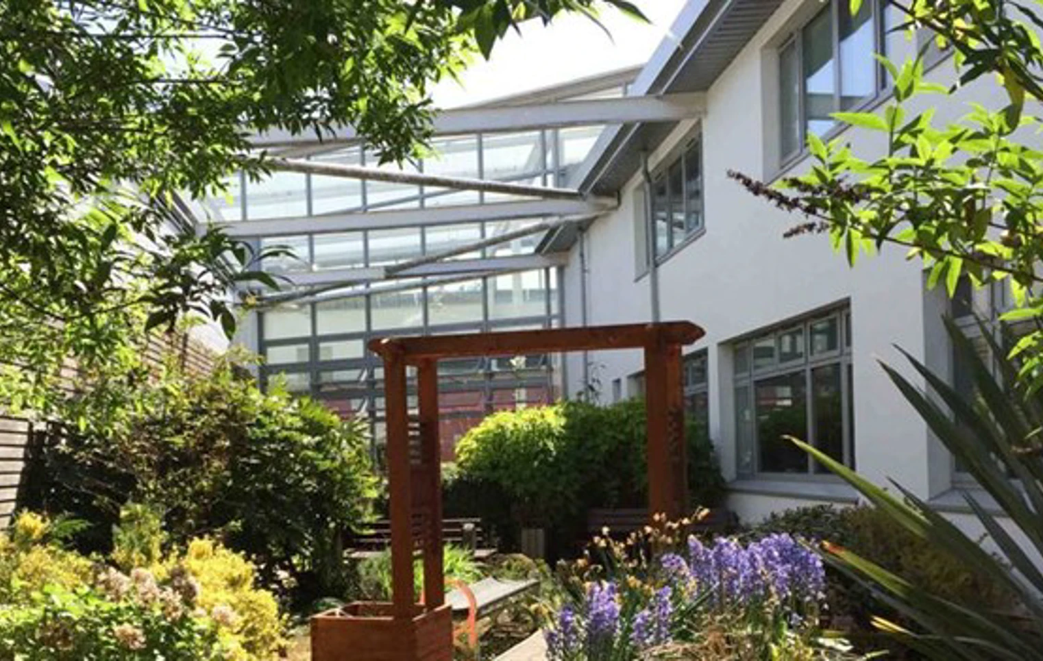 Lush garden courtyard at John Wheatley College featuring flowers, shrubs, and a wooden pergola under a glass canopy. Lush garden courtyard at John Wheatley College featuring flowers, shrubs, and a wooden pergola under a glass canopy.