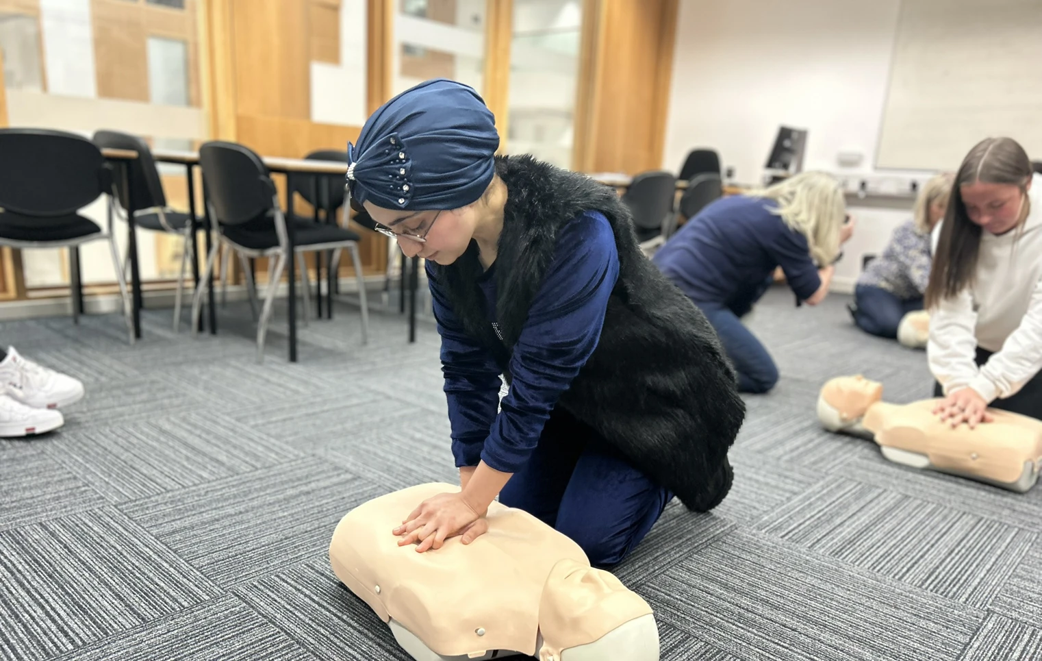 A woman wearing a blue headscarf and glasses practices CPR on a mannequin during a first aid training session in a classroom setting. Other participants are visible in the background. A woman wearing a blue headscarf and glasses practices CPR on a mannequin during a first aid training session in a classroom setting. Other participants are visible in the background.