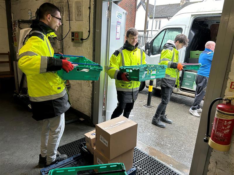 Three young men in high-visibility jackets unloading green crates from a delivery van at a warehouse entrance. Three young men in high-visibility jackets unloading green crates from a delivery van at a warehouse entrance.