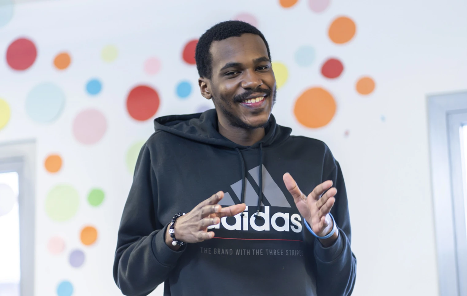 Young man smiling while making expressive hand gestures in front of a colorful polka-dot wall. Young man smiling while making expressive hand gestures in front of a colorful polka-dot wall.