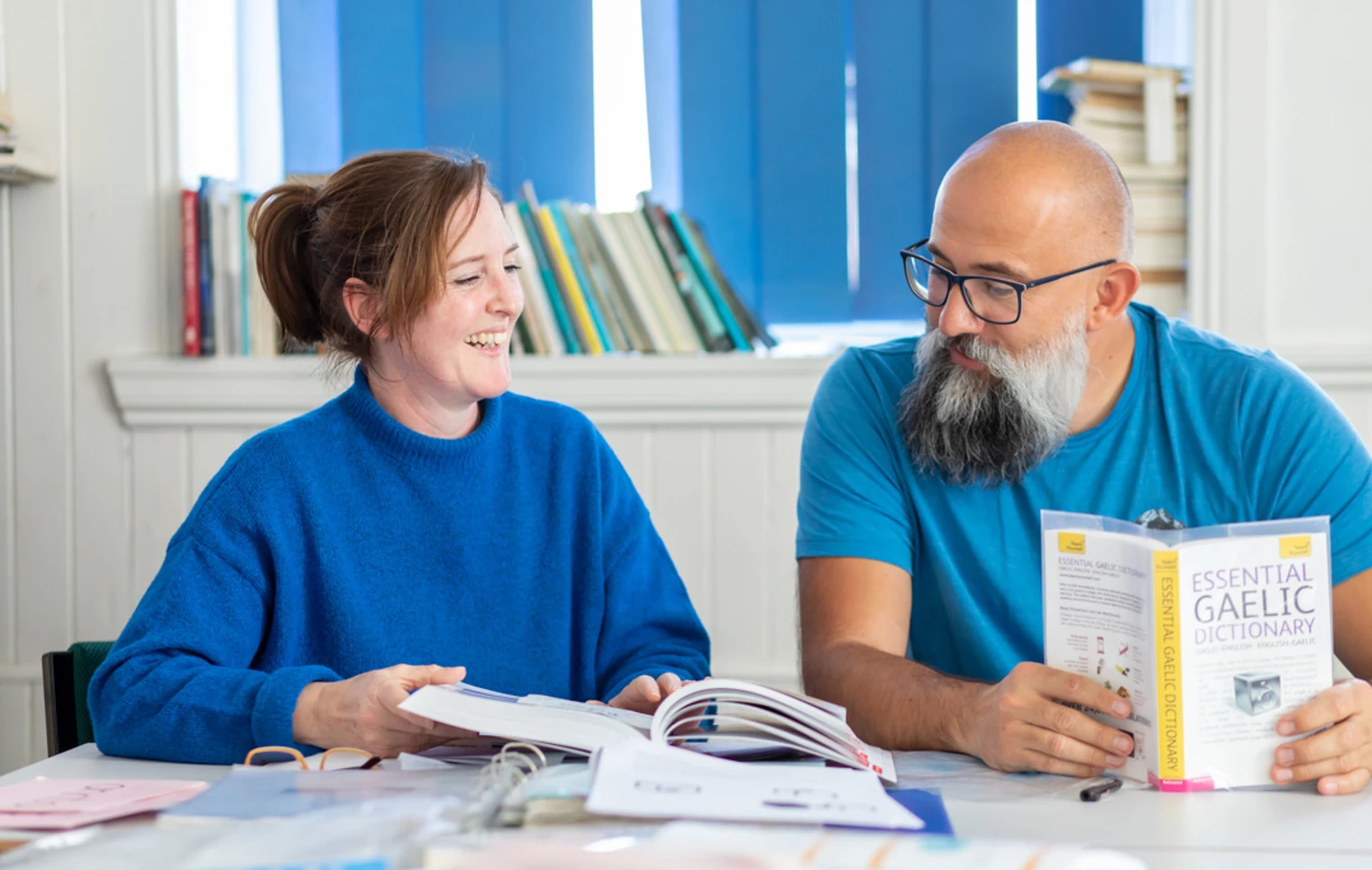 Two students in a Gaelic language class laughing while studying, with a Gaelic-English dictionary open on the desk. Two students in a Gaelic language class laughing while studying, with a Gaelic-English dictionary open on the desk.