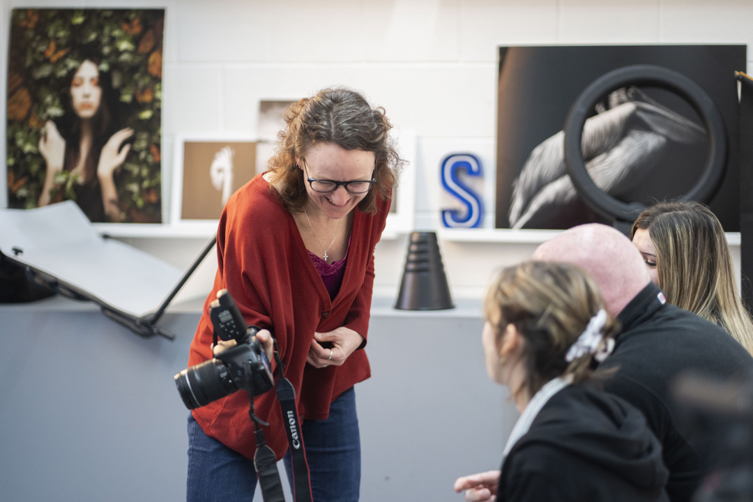 Lecturer smiling and giving feedback to photography students reviewing images on a DSLR camera.
