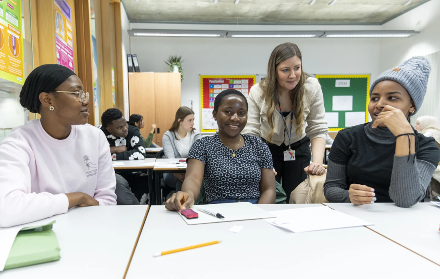 A classroom scene where three female students are smiling and engaging in discussion. A teacher in a beige cardigan leans over the table to interact with them, while other students work in the background. A classroom scene where three female students are smiling and engaging in discussion. A teacher in a beige cardigan leans over the table to interact with them, while other students work in the background.