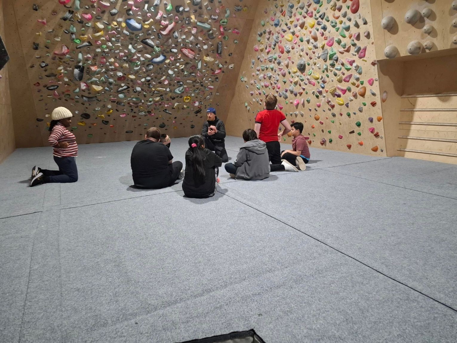 Supported Learning students sitting with an instructor at an indoor climbing centre, listening during a group briefing beneath a bouldering wall.
