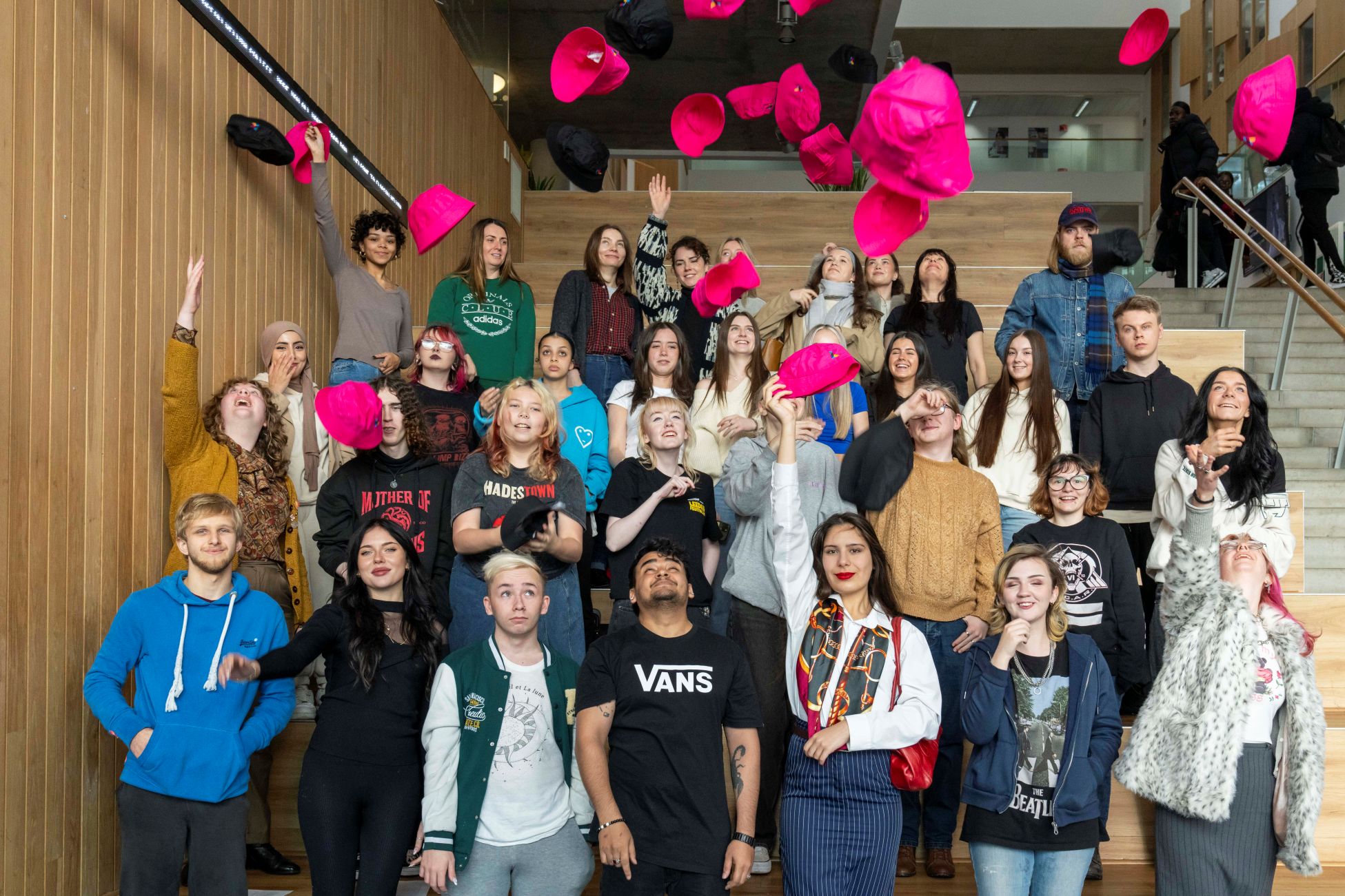 Large group of students gathered on indoor wooden steps, smiling and tossing bright pink and black hats into the air in celebration.
