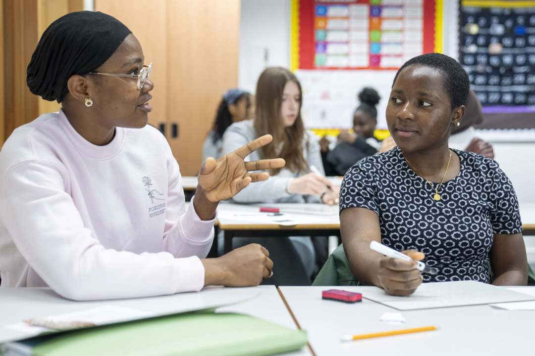 Two female students sit at a desk in a classroom. One student in a pink sweatshirt and a black headscarf is gesturing as she speaks, while the other in a patterned dress listens attentively with a pencil in hand