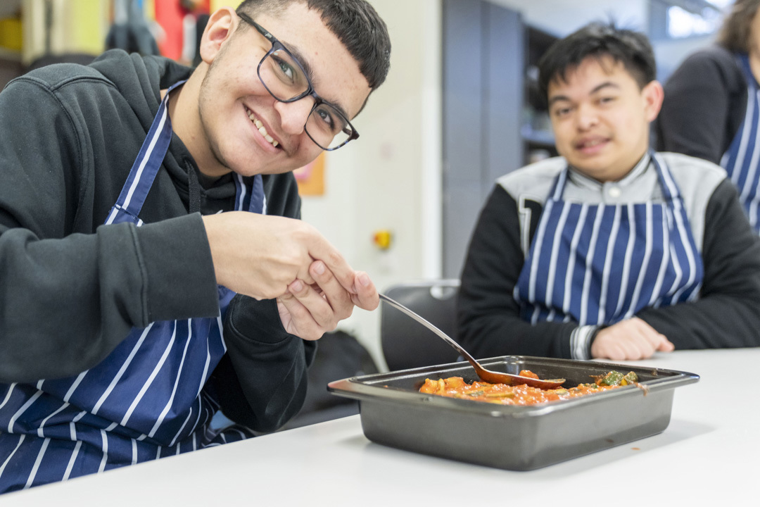 Two young men smiling and preparing a tray of food, wearing striped aprons in a kitchen setting. Two young men smiling and preparing a tray of food, wearing striped aprons in a kitchen setting.