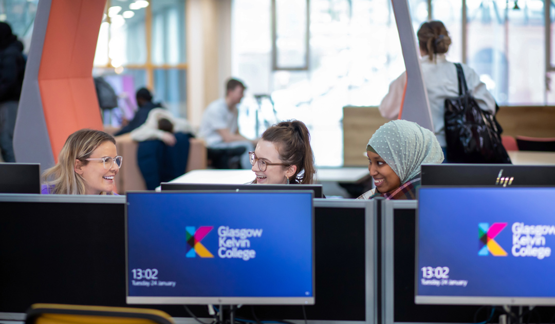 Three female students smiling and chatting while using desktop computers in the library area at Glasgow Kelvin College’s Springburn Campus.