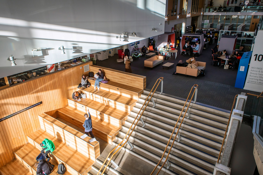 Students sitting and studying in a bright, open-plan learning space at Glasgow Kelvin College Springburn Campus, featuring tiered wooden seating and main stairway.