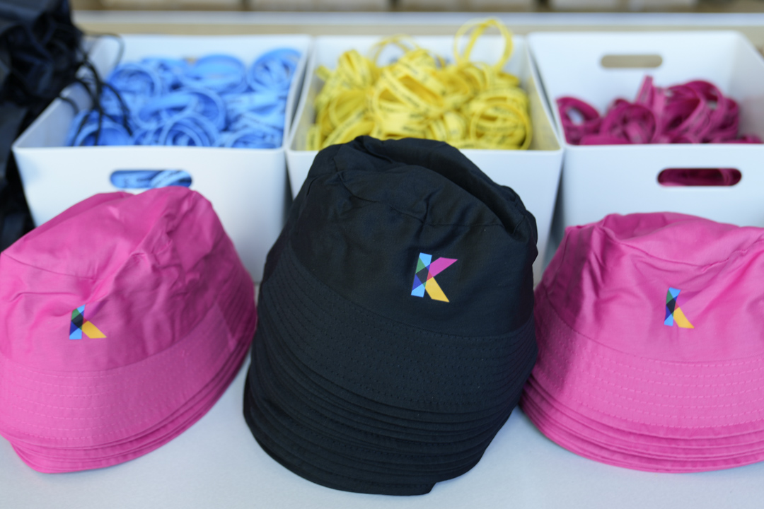Stacked branded bucket hats in pink and black with colourful Kelvin College logos, beside trays of lanyards.