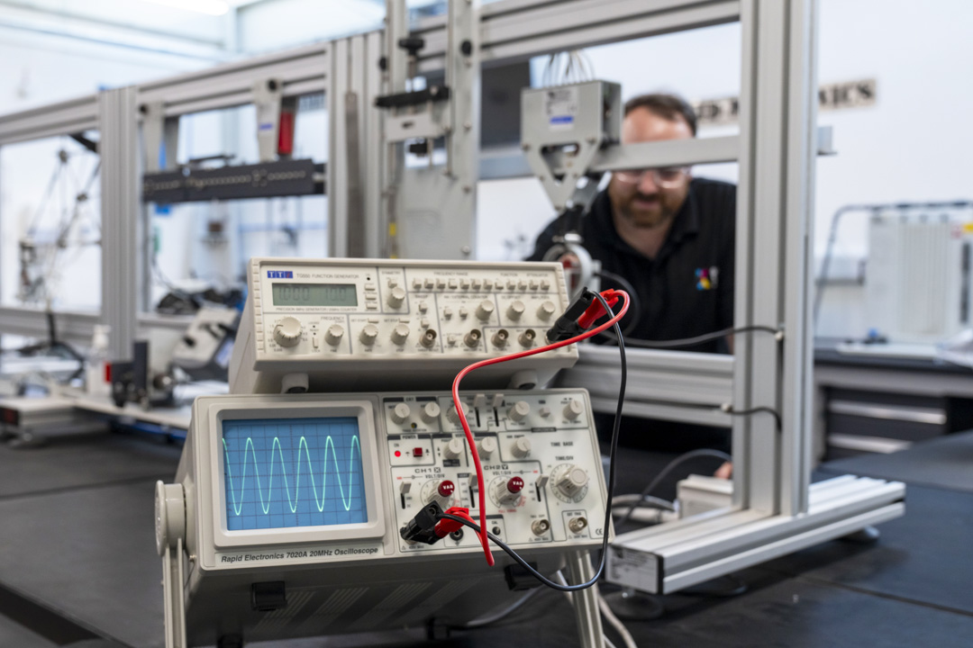 Close-up of an oscilloscope displaying a waveform, connected to a function generator in a laboratory, with a lecturer working in the background.