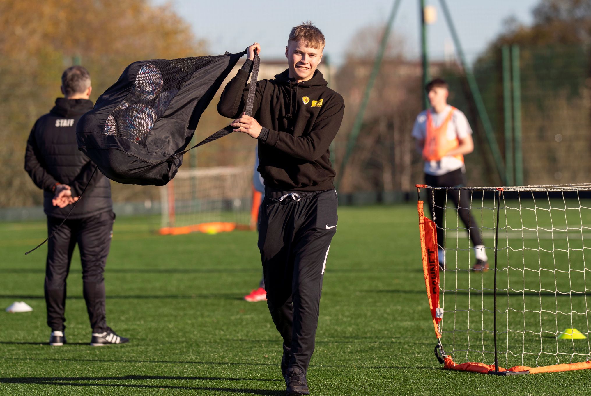 Male student carrying a mesh bag full of footballs across the pitch, smiling during a coaching session.