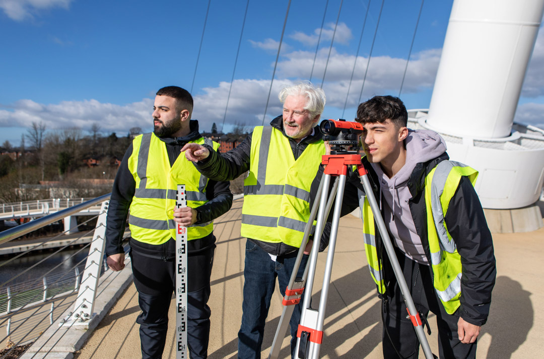 Three individuals wearing high-visibility vests conducting a land surveying exercise on a bridge. One is looking through a theodolite, while another holds a measuring staff, and a lecturer observes under a bright blue sky.