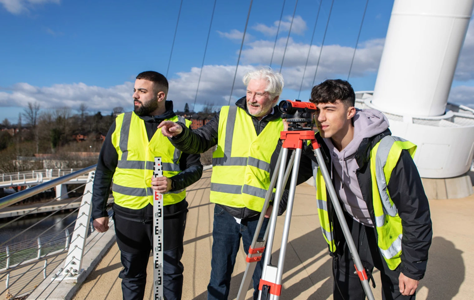Three individuals wearing high-visibility vests conducting a land surveying exercise on a bridge. One is looking through a theodolite, while another holds a measuring staff, and a lecturer observes under a bright blue sky. Three individuals wearing high-visibility vests conducting a land surveying exercise on a bridge. One is looking through a theodolite, while another holds a measuring staff, and a lecturer observes under a bright blue sky.