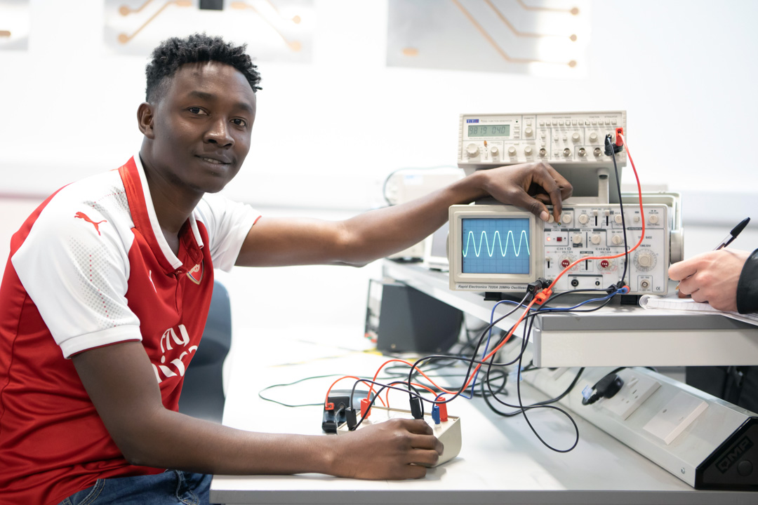 A student in a red football jersey adjusting an oscilloscope in an electronics lab, looking at the camera with a confident expression.