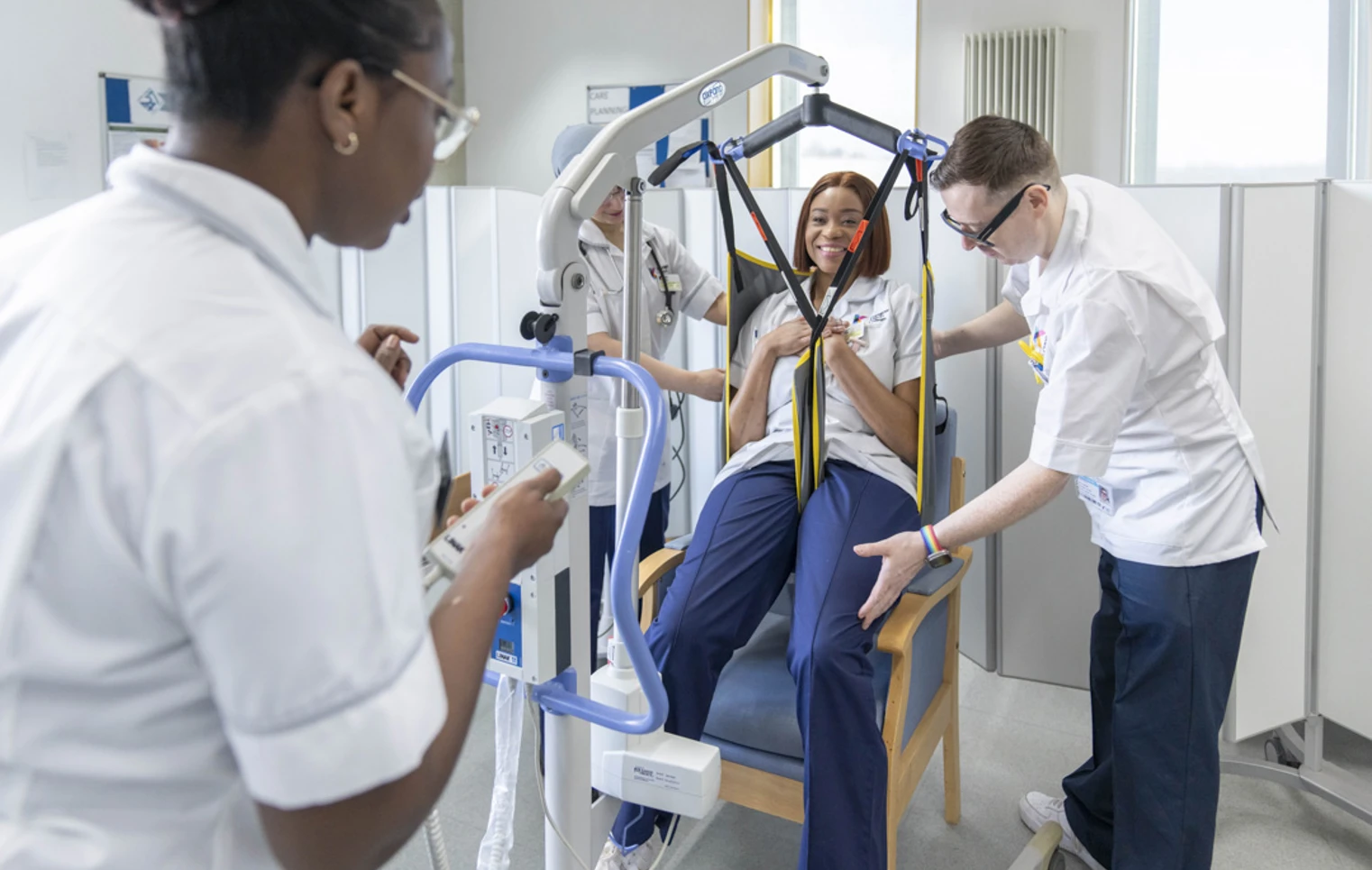 Care students practice using a patient hoist with a seated classmate in a simulated care setting. Care students practice using a patient hoist with a seated classmate in a simulated care setting.