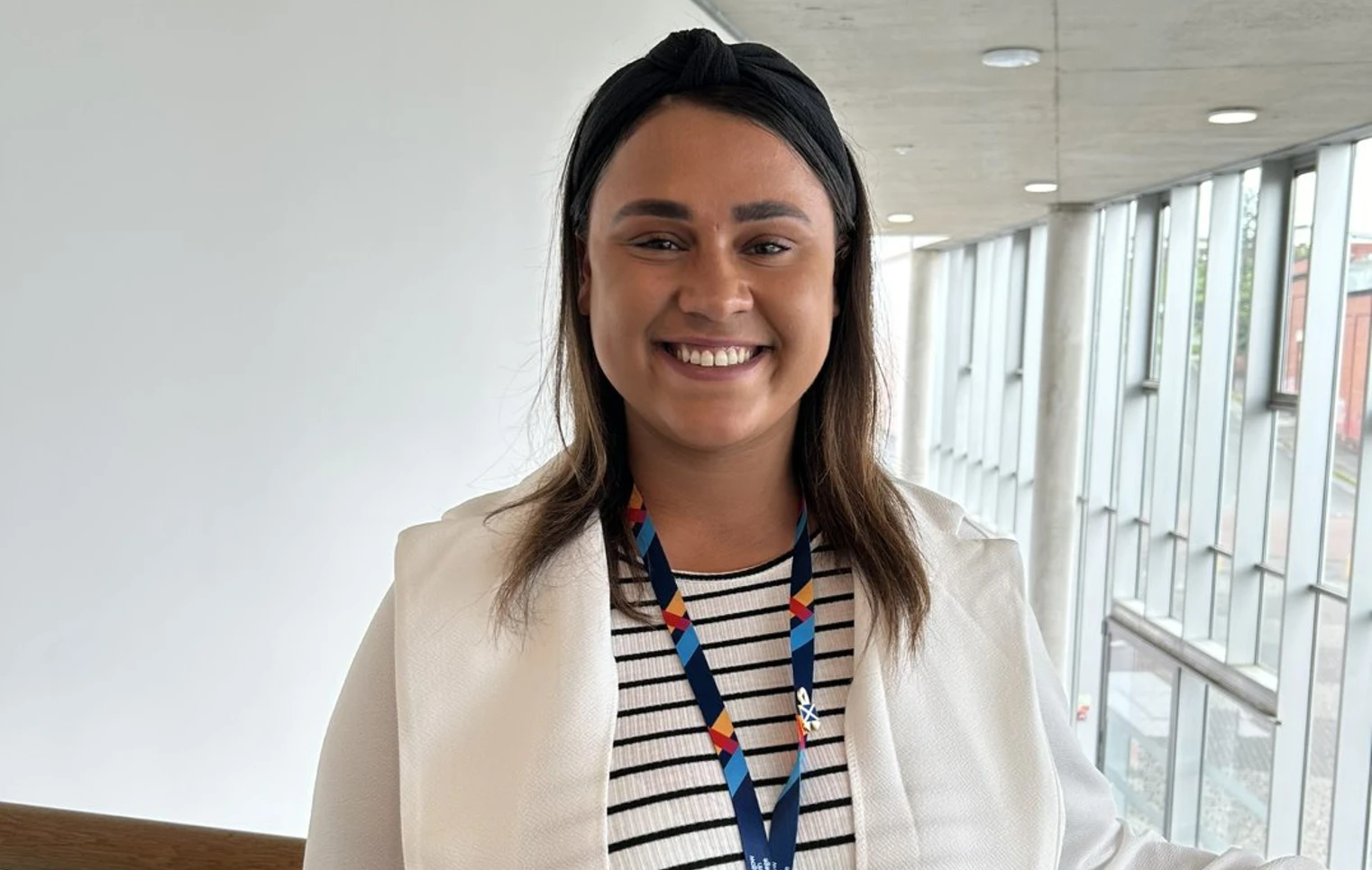 A woman in a white blazer and striped dress stands near a railing in a modern building with large windows. She smiles warmly at the camera, wearing a lanyard with an ID badge. A woman in a white blazer and striped dress stands near a railing in a modern building with large windows. She smiles warmly at the camera, wearing a lanyard with an ID badge.