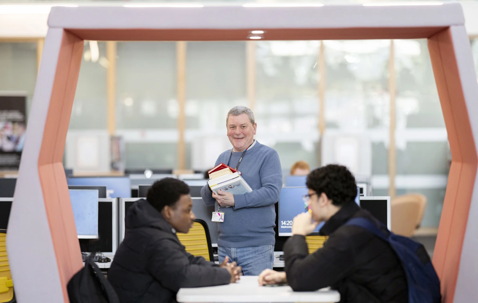 Smiling staff member holding books and standing beside two students in a Glasgow Kelvin College library study pod. Smiling staff member holding books and standing beside two students in a Glasgow Kelvin College library study pod.
